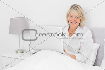 Happy woman sitting in bed with laptop looking at camera