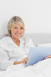 Cheerful woman sitting in bed using tablet pc looking at camera