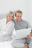 Couple using laptop sitting on bed looking at camera