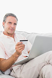 Man on his couch using laptop for shopping online smiling at camera