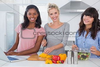 Cheerful friends making salad and using laptop for recipe looking at camera
