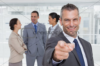 Cheerful businesman pointing at camera with colleagues on background