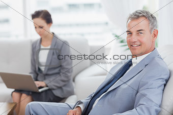 Businessman posing while his colleague working on her laptop