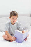 Smiling little boy sitting on bed reading book