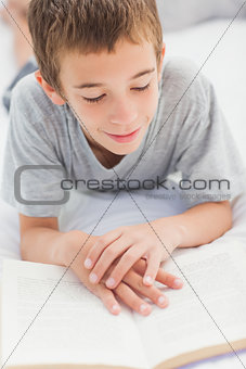 Smiling little boy lying on bed reading book