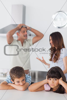 Unhappy siblings sitting in kitchen with their parents who are arguing