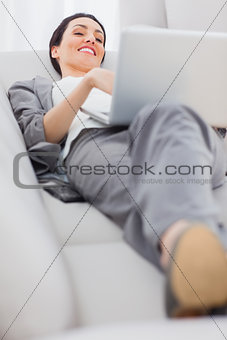 Cheerful businesswoman lying on sofa using laptop