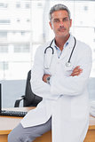 Serious doctor sitting on desk with arms crossed