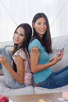 Friends sitting back to back with their tablets smiling at camera