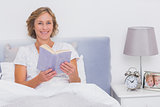 Relaxed blonde woman sitting in bed holding book