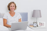Happy blonde woman sitting in bed using laptop