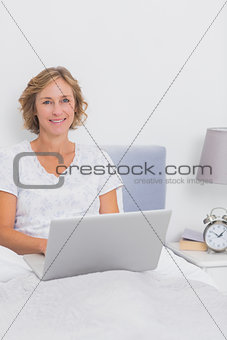 Smiling blonde woman sitting in bed using laptop