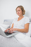 Relaxed blonde woman sitting in bed using laptop