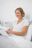 Happy blonde woman sitting in bed using tablet pc