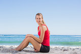 Fit blonde sitting on sand smiling at camera