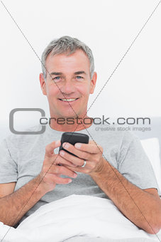 Happy grey haired man sending a text in bed