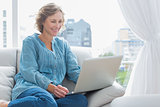 Cheerful blonde woman sitting on her couch using laptop