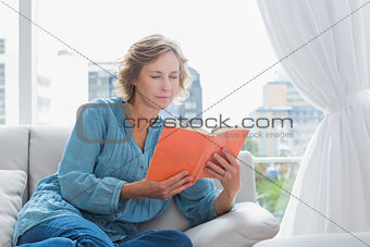 Cheerful blonde woman sitting on her couch reading a book