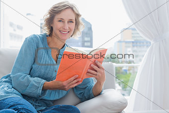 Cheerful blonde woman sitting on her couch holding a book