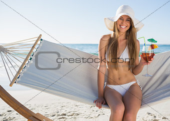 Pretty blonde wearing bikini and sunhat sitting on hammock with cocktail