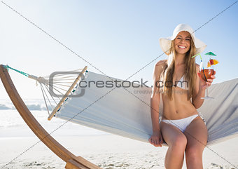 Smiling blonde wearing sunhat sitting on hammock with cocktail