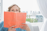 Blonde woman sitting on her couch covering face with orange book