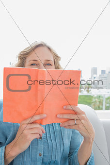 Woman sitting on her couch covering face with orange book