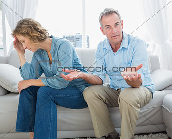 Arguing middle aged couple sitting on the couch with man gesturing at camera