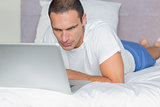 Concentrated man lying on bed using his laptop