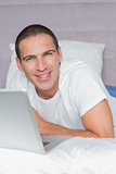 Cheerful man lying on bed using his laptop