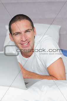 Cheerful man lying on bed using his laptop