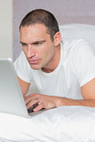 Serious man lying on bed using his laptop