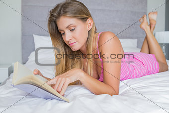 Cheerful blonde lying on her bed reading a book