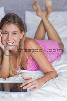 Beautiful blonde lying on her bed using tablet pc smiling at camera