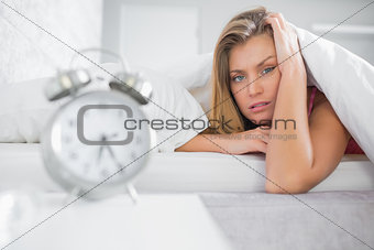 Exhausted blonde looking at camera with alarm clock in foreground