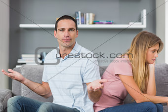 Couple sitting back to back after a fight on the couch with man gesturing at camera