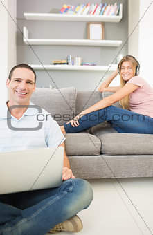 Man sitting on floor using laptop with woman listening to music on the sofa both smiling at camera