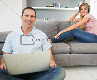 Man sitting on floor using laptop with woman listening to music on the sofa both looking at camera