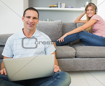 Man sitting on floor with laptop with woman listening to music on the sofa both looking at camera