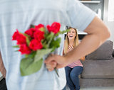 Man hiding bouquet of roses from smiling girlfriend on the couch