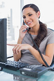 Smiling secretary answering land line and pointing at computer screen