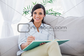 Smiling gorgeous brunette writing while sitting on the sofa