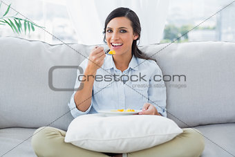 Cheerful woman sitting on the couch crossing legs eating fruits