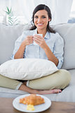 Cheerful woman on sofa drinking coffee and having croissant