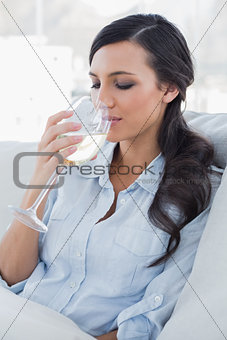 Relaxed pretty brunette drinking white wine sitting on sofa