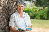 Cheerful mature woman holding book sitting on tree trunk