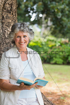 Smiling mature woman reading book leaning on tree trunk