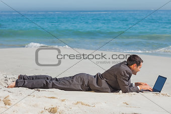 Young businessman lying on the sand typing on his laptop