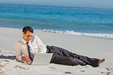 Cheerful young businessman lying on the sand with his laptop