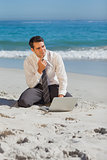 Pensive young businessman sitting on the sand with his laptop